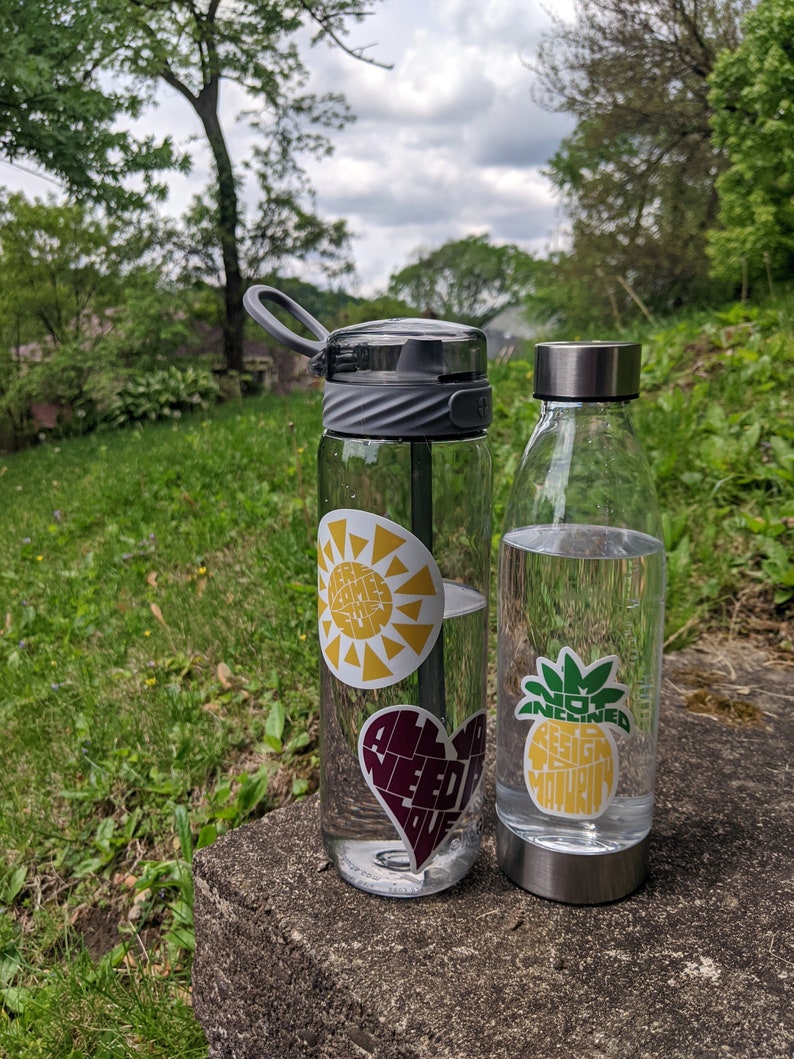 Puede incluir: Dos botellas de agua con pegatinas coloridas. La botella de la izquierda tiene una pegatina de sol amarilla con el texto "Feel Some Sun", una pegatina de coraz&oacute;n morado con el texto "All You Need Is Love", y una pegatina en blanco y negro con un dise&ntilde;o de sol y luna. La botella de la derecha tiene una pegatina de pi&ntilde;a verde y amarilla con el texto "Not Inclined To Design Maturity".