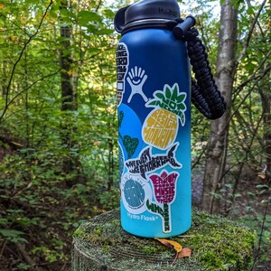 Puede incluir: Una botella de agua azul Hydro Flask con una correa de transporte negra. La botella est&aacute; cubierta de pegatinas coloridas con varios dise&ntilde;os y textos, incluyendo una pi&ntilde;a, un tibur&oacute;n y una flor.