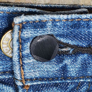 May include: Close-up of blue denim jeans with a button and buttonhole. The button is gold-colored with the text "Levi's" and the buttonhole is black. The denim fabric is textured and shows signs of wear.