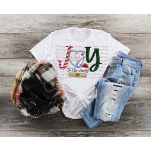 Puede incluir: Camiseta blanca con un dise&ntilde;o de "Joy to the world" en verde, rojo y azul que incluye un estetoscopio, un bast&oacute;n de caramelo y un coraz&oacute;n con un vendaje. El dise&ntilde;o est&aacute; en una camiseta blanca con un fondo blanco.