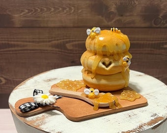 May include: A white wooden pedestal with a round top and three legs. On top of the pedestal is a small wooden cutting board with a honey dipper and a stack of honey-drizzled donuts decorated with bees.