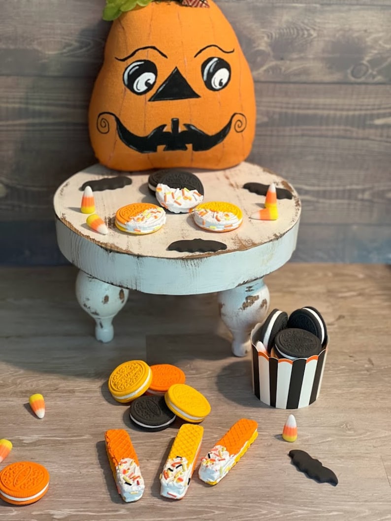 May include: A Halloween-themed table setting with a pumpkin-shaped cushion, black bats, and orange and black biscuits. There are also candy corn and a small white table with three legs.