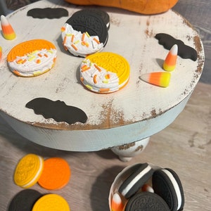 May include: A white tiered cake stand with a blue base, decorated with black bats, orange and yellow candy corn, and Halloween-themed cookies. The cookies are decorated with white icing and orange and yellow sprinkles.
