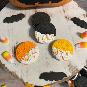 May include: Three Halloween-themed Oreo cookies decorated with white icing and orange and yellow sprinkles. The cookies are on a white wooden surface with black bats and candy corn.