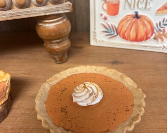 May include: A small pumpkin pie with a swirl of whipped cream on top. The pie is in a brown pie crust and is sitting on a wooden table. The background includes a wooden shelf with a sign that says "Pumpkin Spice and everything nice."
