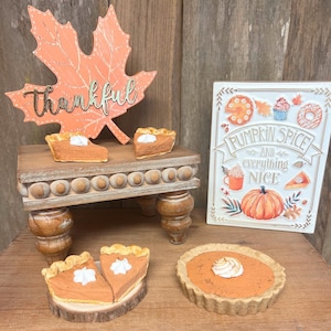 May include: A wooden table with a fall-themed sign that says "Pumpkin Spice and Everything Nice". There are four miniature pumpkin pies on the table and a wooden leaf with the word "thankful" written on it.