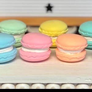 May include: A collection of pastel-colored macarons, each with a white filling. The macarons are arranged in a row on a white surface.