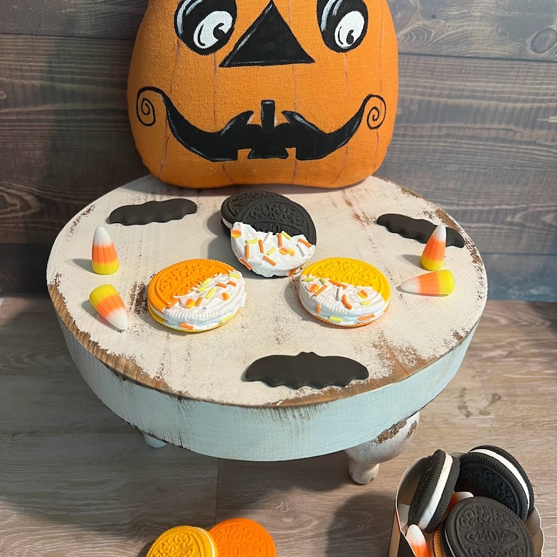May include: A white pedestal stand with three legs, decorated with black bats and orange and yellow candy corn. The stand is topped with four cookies, two with black and white frosting and two with orange and white frosting. A pumpkin-shaped cushion sits on the floor behind the stand.
