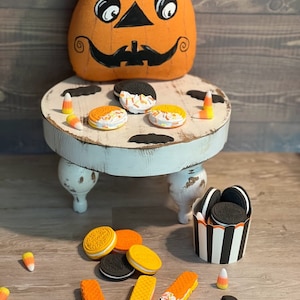 May include: A Halloween-themed table setting with a pumpkin-shaped cushion, black bats, and orange and black biscuits. There are also candy corn and a small white table with three legs.