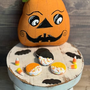 May include: A white painted wooden stool with three legs, decorated with black bats and orange and white candy corn.  On top of the stool are three orange and white cookies with sprinkles, and one black and white cookie.  A smiling orange pumpkin with a black face is sitting on the floor behind the stool.