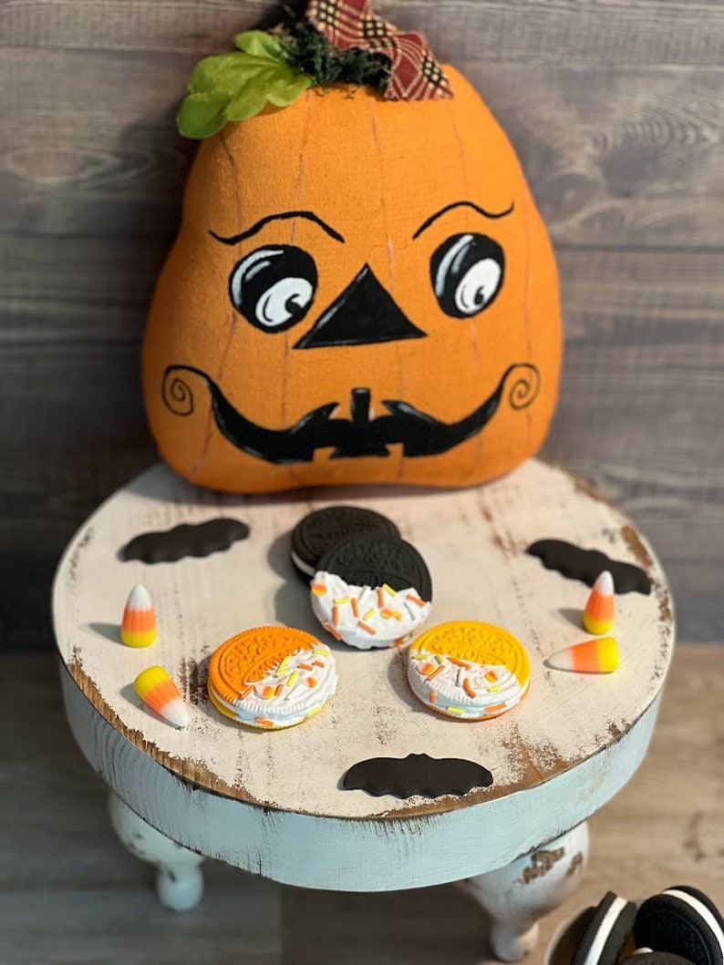 May include: A white painted wooden stool with three legs, decorated with black bats and orange and white candy corn.  On top of the stool are three orange and white cookies with sprinkles, and one black and white cookie.  A smiling orange pumpkin with a black face is sitting on the floor behind the stool.