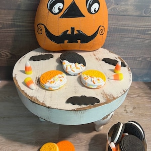 May include: A white pedestal stand with three legs, decorated with black bats and orange and yellow candy corn. The stand is topped with four cookies, two with black and white frosting and two with orange and white frosting. A pumpkin-shaped cushion sits on the floor behind the stand.