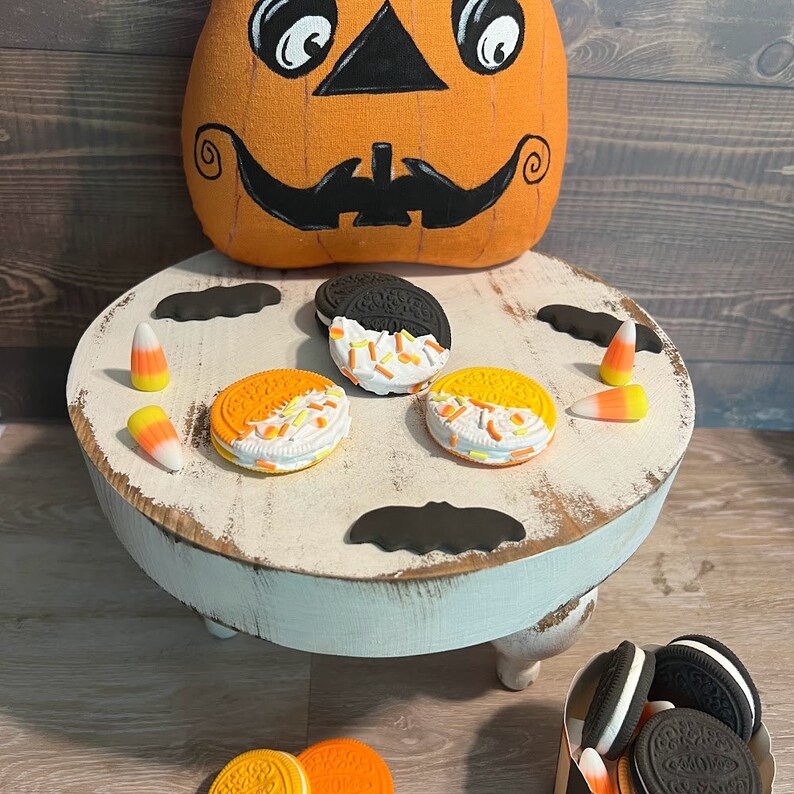 May include: A white pedestal stand with three legs, decorated with black bats and orange and yellow candy corn. The stand is topped with four cookies, two with black and white frosting and two with orange and white frosting. A pumpkin-shaped cushion sits on the floor behind the stand.