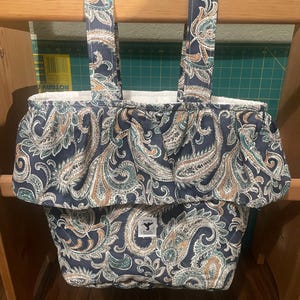 May include: A navy blue fabric tote bag featuring a white, teal, and tan paisley pattern. The bag has two top handles, a ruffled top edge, and a white interior lining. A small rectangular label with a hummingbird logo is centered on the front.