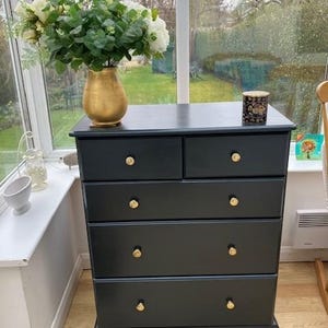 May include: A dark grey chest of drawers with five drawers and gold-coloured knobs. A gold vase with white flowers and greenery sits on top, along with a small decorative container. The chest of drawers is in a room with a large window.