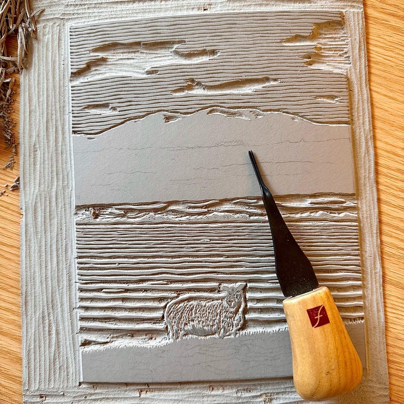 May include: A gray linocut print depicting a sheep in a landscape with a mountain range and a tool with a wooden handle. The linocut is on a light gray surface. The tool has a black blade and a wooden handle with a red square logo.