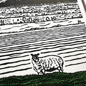 May include: Black and white linocut print of a sheep standing on green grass, with a landscape background. The landscape features a mountain range, fields, and trees. The sky is white with a few clouds.