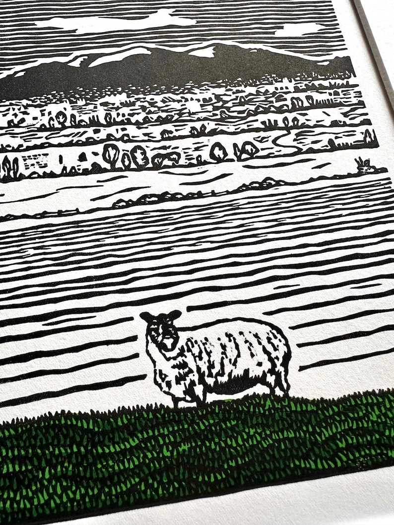 May include: Black and white linocut print of a sheep standing on green grass, with a landscape background. The landscape features a mountain range, fields, and trees. The sky is white with a few clouds.