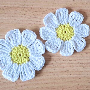 May include: Two crocheted white daisy flowers with yellow centres. The flowers are on a light brown wooden surface.