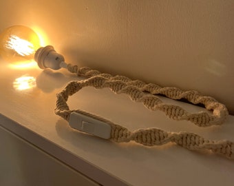 Op de afbeelding: Een decoratieve macramé lamp met een warme, gloeiende gloeilamp. De lamp heeft een witte lamphouder en een geweven, crèmekleurig macramé snoer met een inline schakelaar. De lamp staat op een wit oppervlak.
