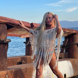 May include: A woman with long blonde braids wears a white crochet cover-up over a blue and white striped bikini. She stands on a rusty metal platform with a wooden plank overhead. The sea is visible in the background.