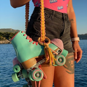 May include: A pair of mint green quad roller skates with pink laces, suspended by a handmade yellow and pink macrame strap featuring a wooden ring and a small tassel. The skates have matching mint green wheels and pink crochet toe caps.