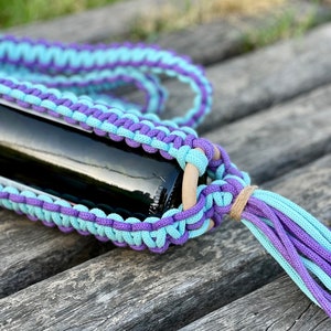 May include: A crocheted bottle holder made with light blue and purple yarn. The holder is wrapped around a wine bottle and has a tassel on the end.