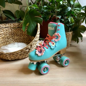 Op de afbeelding: Een paar turquoise blauwe rollerskates met witte wielen en een roze en wit gecrochet bloemendesign. De skates hebben het woord "Impala" aan de zijkant gedrukt.