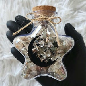 May include: A star-shaped clear glass jar filled with small, polished, translucent stones. The jar features a black crescent moon and star decal on the front, a cork stopper, and a decorative brown twine bow tied around the neck of the bottle.