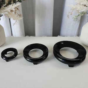 May include: Three black, glossy sphere stands of varying sizes are arranged on a white surface. Each stand has a circular opening in the center and a small, rectangular base. Decorative flowers and vases are in the background.