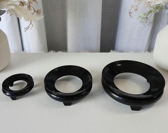 May include: Three black, glossy sphere stands of varying sizes are arranged on a white surface. Each stand has a circular opening in the center and a small, rectangular base. Decorative flowers and vases are in the background.