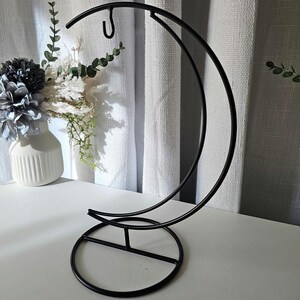 May include: A black metal display stand featuring a crescent moon shape with a circular base and a small hook at the top for hanging items. The structure is made of thin, curved metal rods, providing a stable frame for decorative objects.
