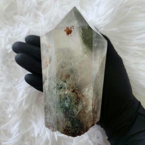 May include: A clear, hexagonal quartz crystal point featuring internal green chlorite inclusions and small reddish-brown mineral deposits. The crystal has a polished, multi-faceted surface with a pointed top and a rough, uneven base.