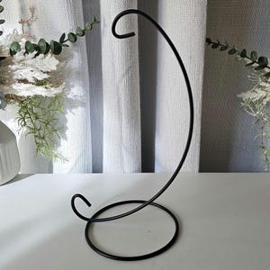 May include: A black metal display stand featuring a circular base and a curved, C-shaped arm with a hook at the top, designed for hanging small decorative items. The stand is constructed from thin, smooth wire with a matte finish.