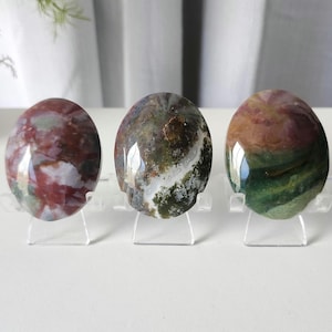 May include: Three polished oval stones displayed on clear plastic stands. The left stone features mottled red, white, and green patterns. The center stone shows dark green, brown, and white inclusions. The right stone displays blended green, brown, and red hues.