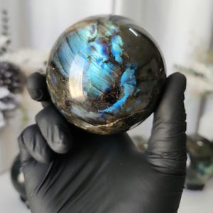 May include: A polished labradorite sphere, held in a gloved hand. The sphere displays iridescent blue and green flashes across a dark, speckled surface. The gemstone is a natural, decorative item.