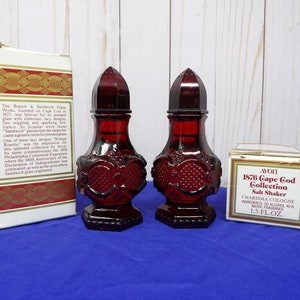 May include: Two red glass salt shakers with a detailed design, sitting on a blue surface. The salt shakers are in their original boxes, which are labeled "Avon 1876 Cape Cod Collection Salt Shaker".