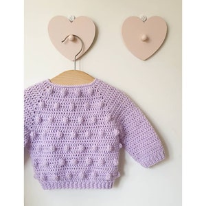 Puede incluir: Un suéter de crochet lavanda con un patrón de punto de textura de pompón. El suéter está colgado en una percha de madera con dos ganchos en forma de corazón rosa en la pared detrás.