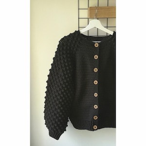 May include: A black crocheted cardigan with a textured, bobbly design on the sleeves. The cardigan has a round neckline and is fastened with wooden buttons.