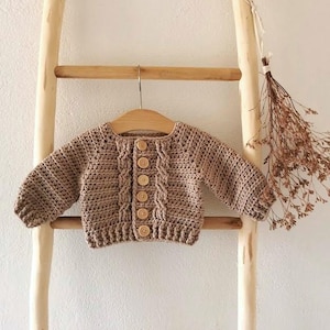 May include: A brown crocheted cardigan with wooden buttons. The cardigan is hanging on a wooden ladder.