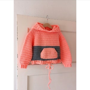 May include: A coral pink crocheted hooded sweater with a grey stripe and a matching pocket. The sweater has a drawstring at the bottom.