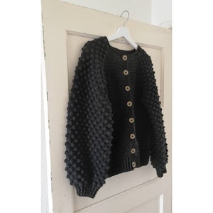 Op de afbeelding: Een zwarte gebreide cardigan met een textuur, bobbelig ontwerp. De cardigan heeft een knoopsluiting en lange mouwen.