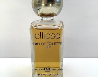 May include: A vintage perfume bottle of "Ellipse" Eau de Toilette. The clear, rectangular bottle contains a golden-colored liquid and is topped with a silver spherical cap. The label reads "JACQUES FATH PARFUMS PARIS" and "57ml 2 fl. oz."
