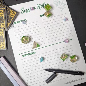 May include: A white sheet of paper with green lines and a title that says "Session Notes". There are two columns labeled "Objectives" and "NPC". The bottom of the page has a line and the word "Happenings" written below it. There are several dice scattered on the page, including green, blue, and pink dice.