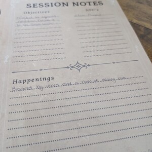 May include: A tan-colored session notes sheet with the title "MONSTERS MET / SESSION NOTES" at the top. The sheet includes sections for objectives, NPC's, and happenings, with lines for writing. The text "Insared...by...vines...and...a...rush...of...heavy...ram..." is handwritten.