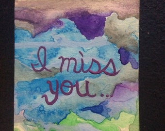 Könnte beinhalten: Aquarellmalerei eines bewölkten Himmels in Blautönen, Grün, Lila und Grau mit den Worten "I miss you..." in Lila.