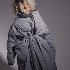 Op de afbeelding: Een grijze pufferjas met een hoge kraag en een strikceintuur. De jas heeft grote zakken en een volumineus ontwerp. Het model draagt de jas, met het hoofd naar beneden. De achtergrond is effen grijs.