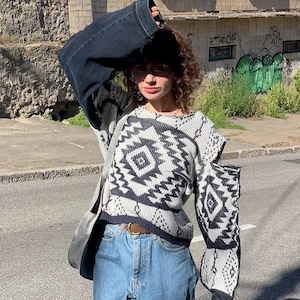 Puede incluir: Una persona con un suéter gris y negro estampado, jeans azules y zuecos blancos. El suéter tiene un diseño geométrico. La persona también lleva una chaqueta vaquera negra y un bolso de hombro gris. El atuendo es casual y elegante.