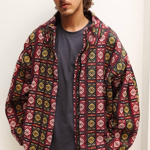 May include: A zipped jacket with a geometric pattern in red, yellow, and white on a dark background. The jacket is worn over a grey t-shirt and beige trousers. The style is casual and contemporary.