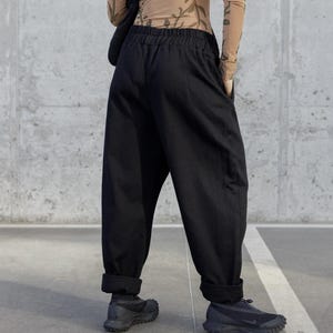 Op de afbeelding: Zwarte, wijde broek met omgeslagen zomen. De outfit wordt gecombineerd met zwarte sneakers en een doorschijnende top met lange mouwen en een natuurpatroon. De set staat tegen een betonnen achtergrond.